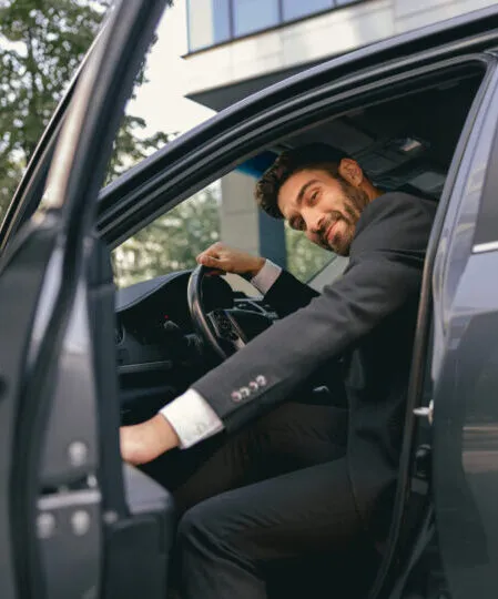 Smiling businessman in suit looking at camera while sitting in driver seat with opened door